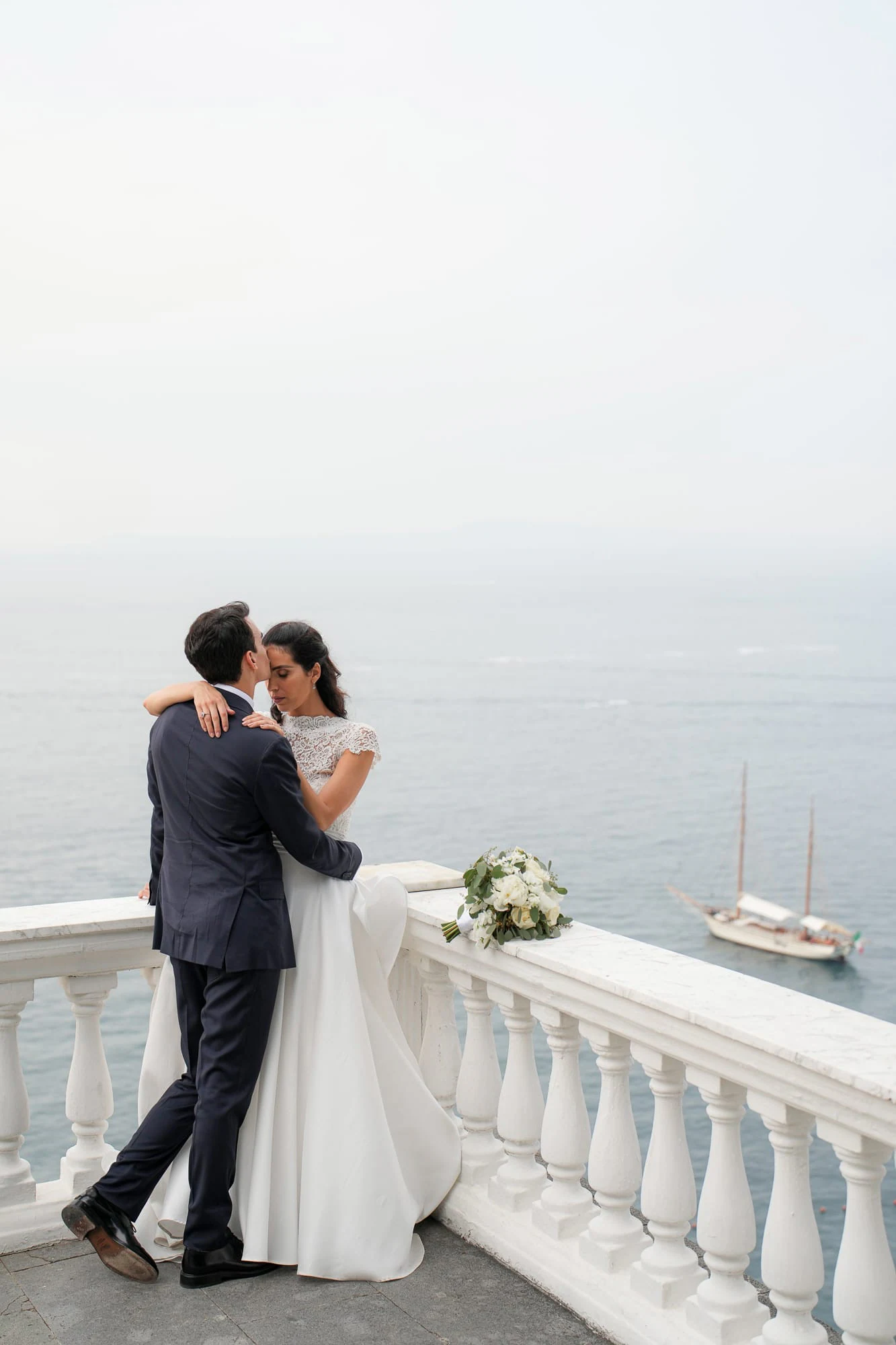 Bride and groom embracing at the white balustrade of La Cocumella above the sea, a sailing boat below.