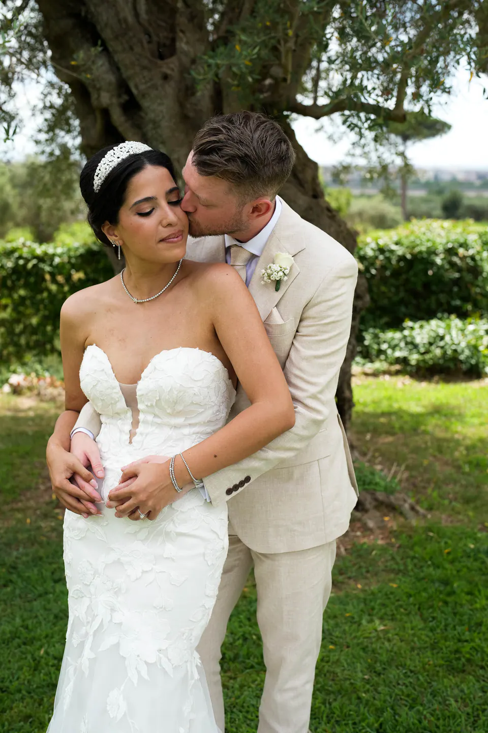 Le marié enlace la mariée par derrière et l’embrasse sur la joue dans le jardin.