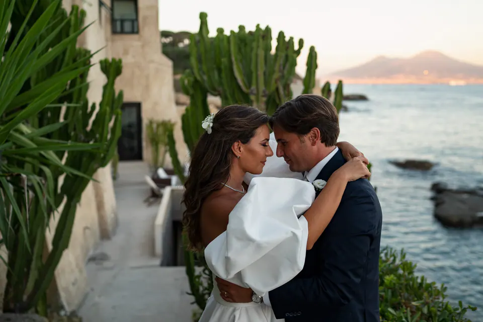 Groom embracing the bride among the cacti of the seaside garden, Vesuvius in the distance.