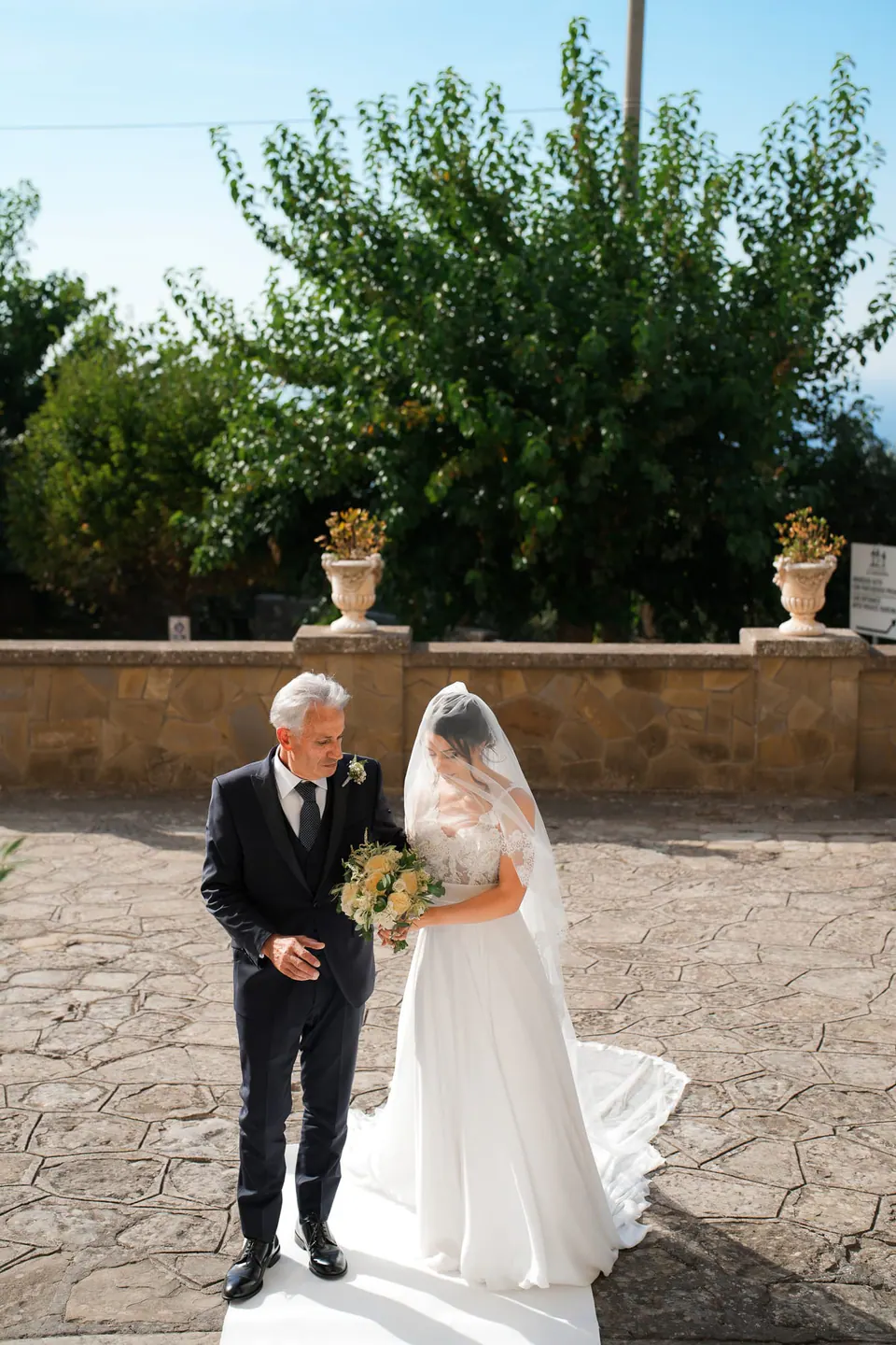Father walking the bride across the stone courtyard to the church.