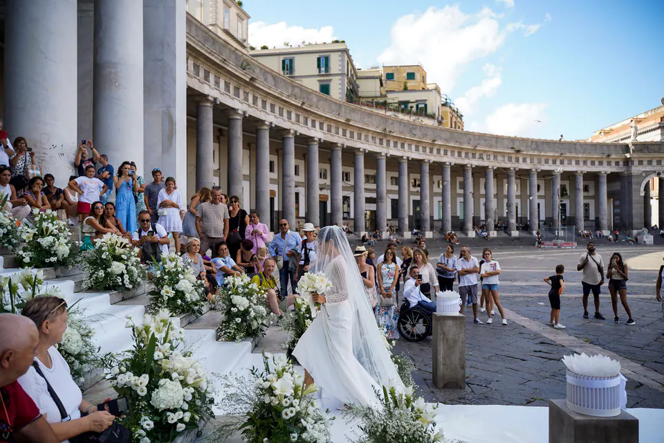 Guests seated among white flowers beneath the colonnade of Piazza del Plebiscito, the bride arriving.