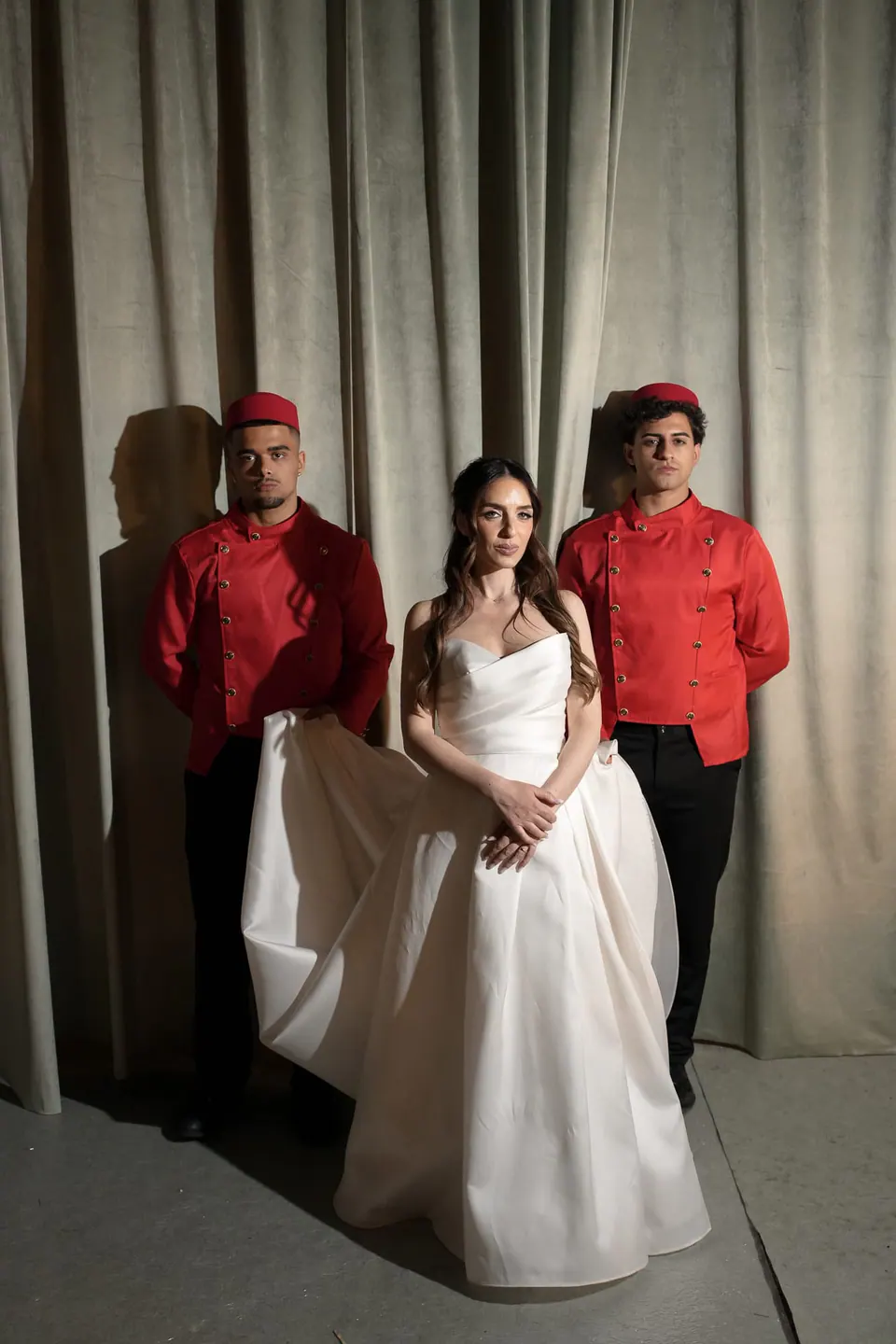 La novia entre dos camareros con chaqueta roja delante de una cortina.
