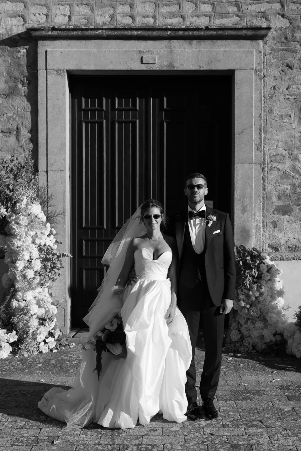 Ritratto in bianco e nero degli sposi con gli occhiali da sole davanti alla porta della chiesa.