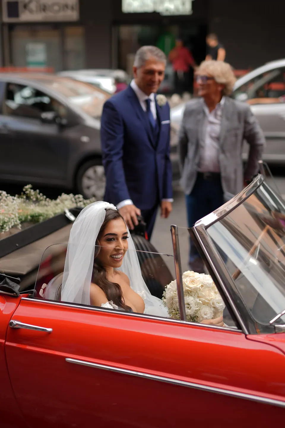 Bride seated in the red vintage convertible, her veil around her, family standing beside the car.