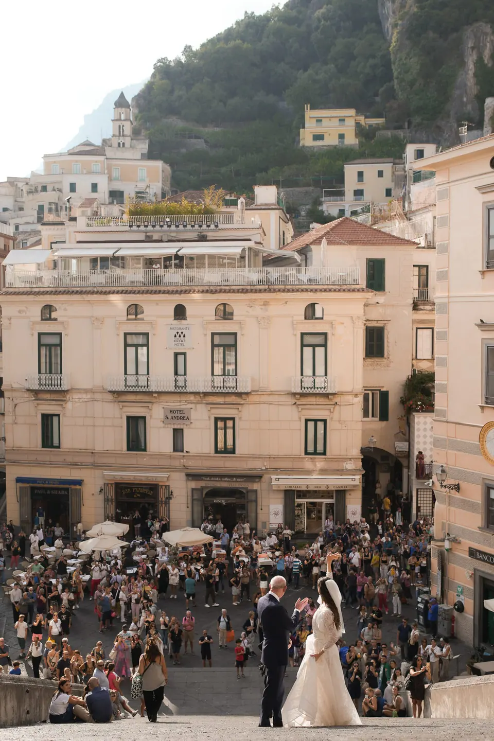 Bride and groom in the crowded Piazza Duomo, Amalfi and its hills behind them.