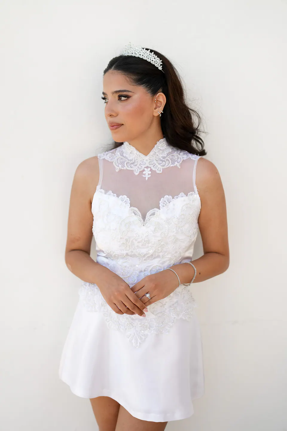 La mariée en robe courte de dentelle blanche et diadème contre un mur blanc, le regard de côté.