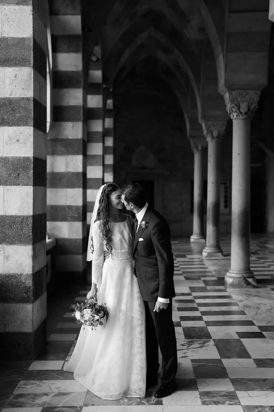 Black and white photo of the couple beneath the striped arches of the Amalfi Cathedral atrium.