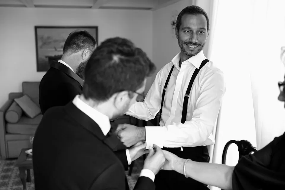 Black and white photo of the groom getting dressed as his friends help with his shirt.