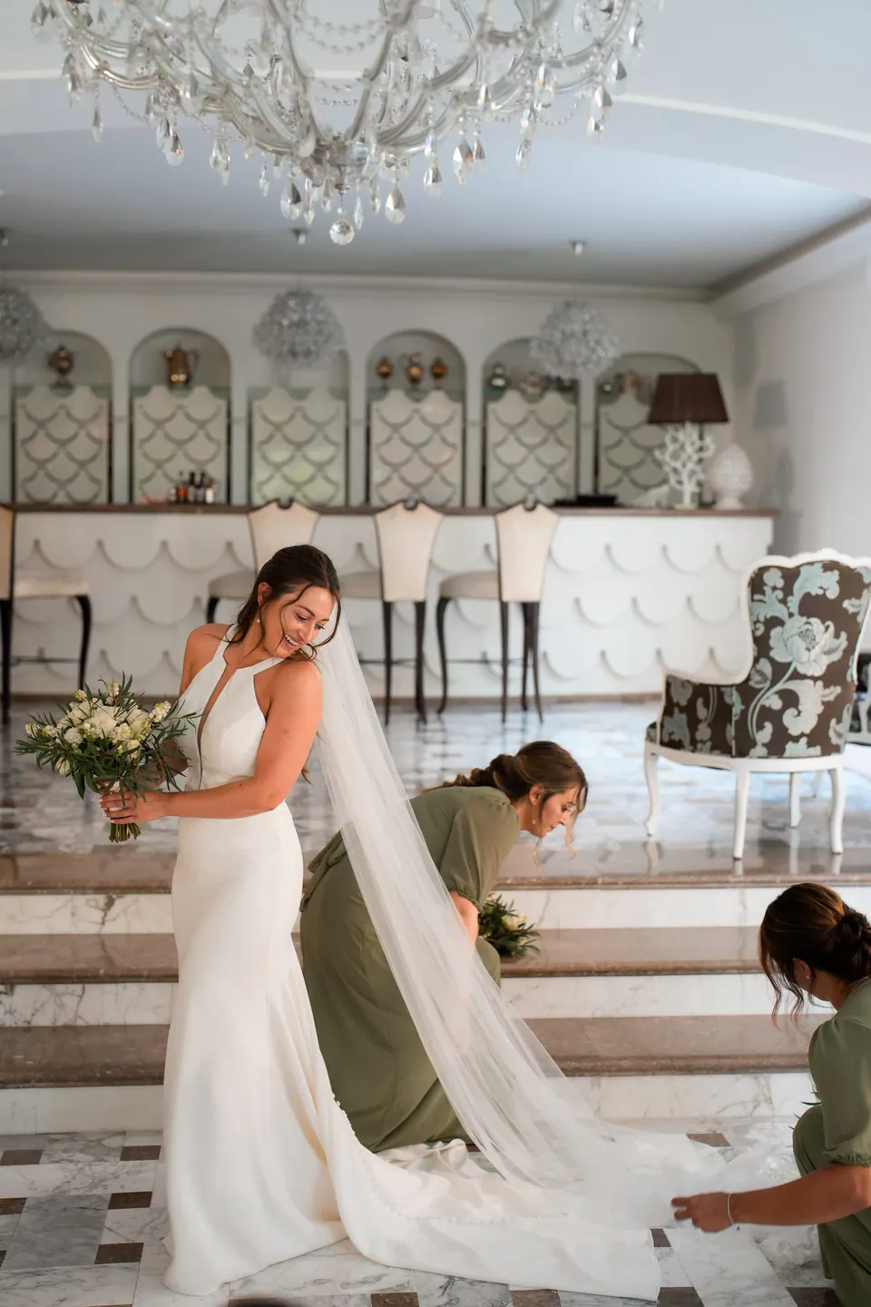 Bride in the chandelier-lit salon of Relais Blu, a bridesmaid arranging her long veil.
