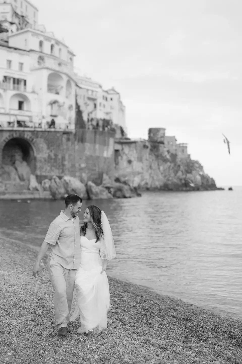 Black and white photo of the couple walking along the shore below the cliffs of Amalfi.