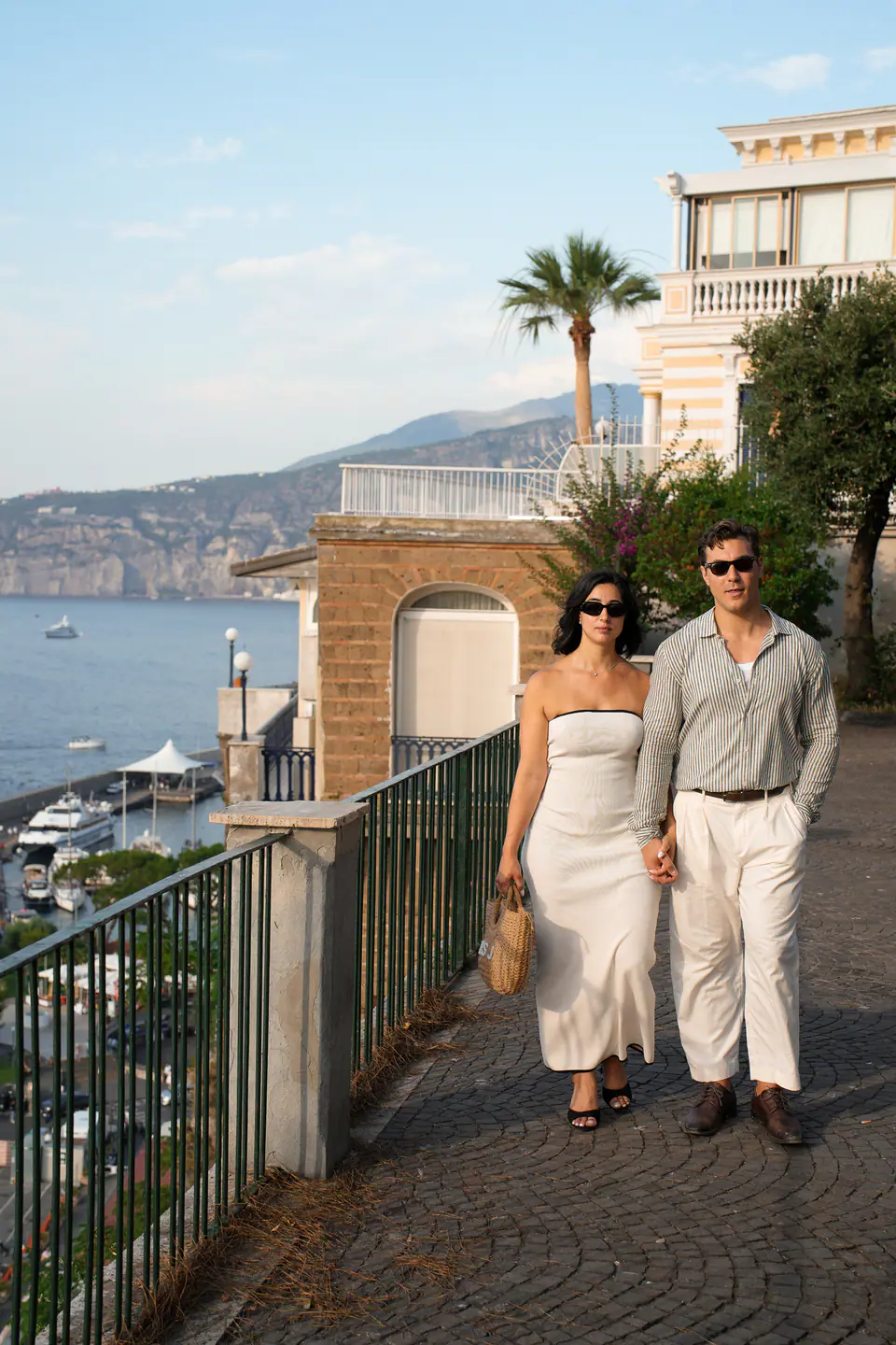 Couple walking along a terrace above the sea, palm trees behind them.