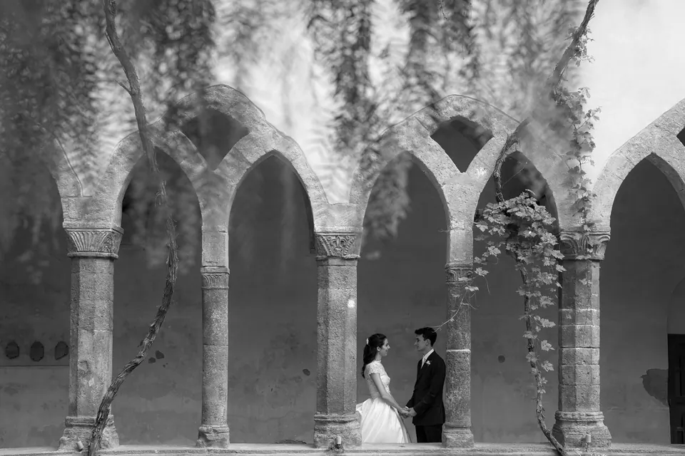 Black and white photo of bride and groom small beneath the arches of the cloister