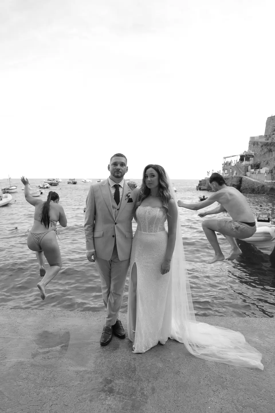 Black and white photo of the couple standing by the sea while swimmers dive from the rocks.