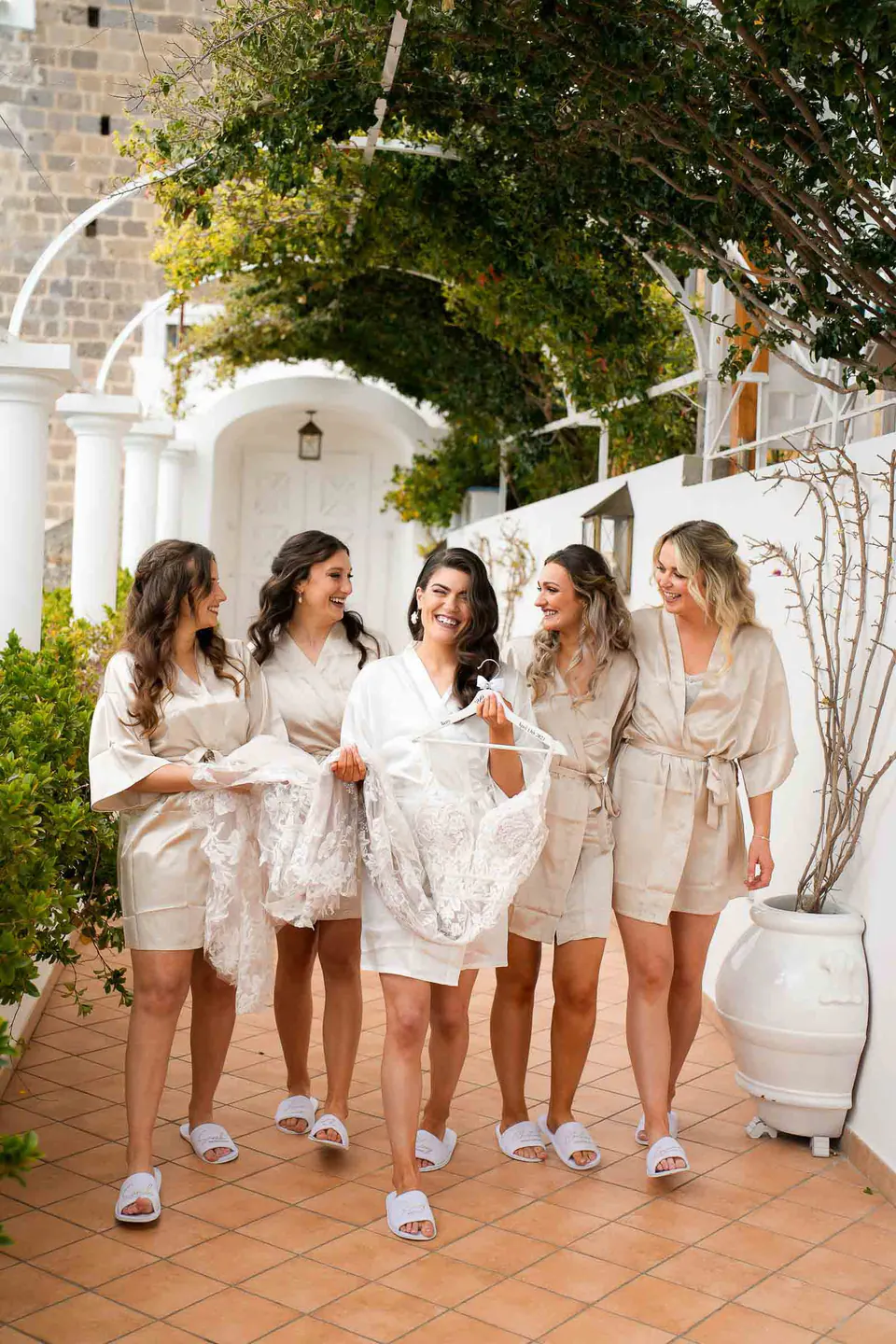 La sposa e le damigelle in abiti coordinati che camminano insieme, risate e sole mattutino.
