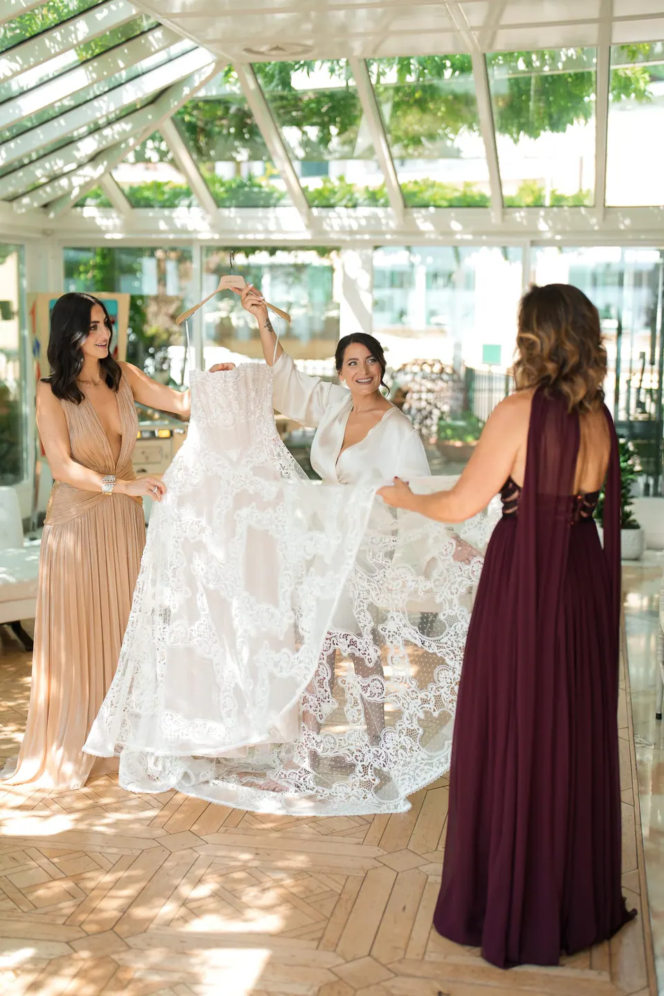 Brautjungfern in Champagner und Bordeaux halten in der Veranda das Spitzenkleid hoch.