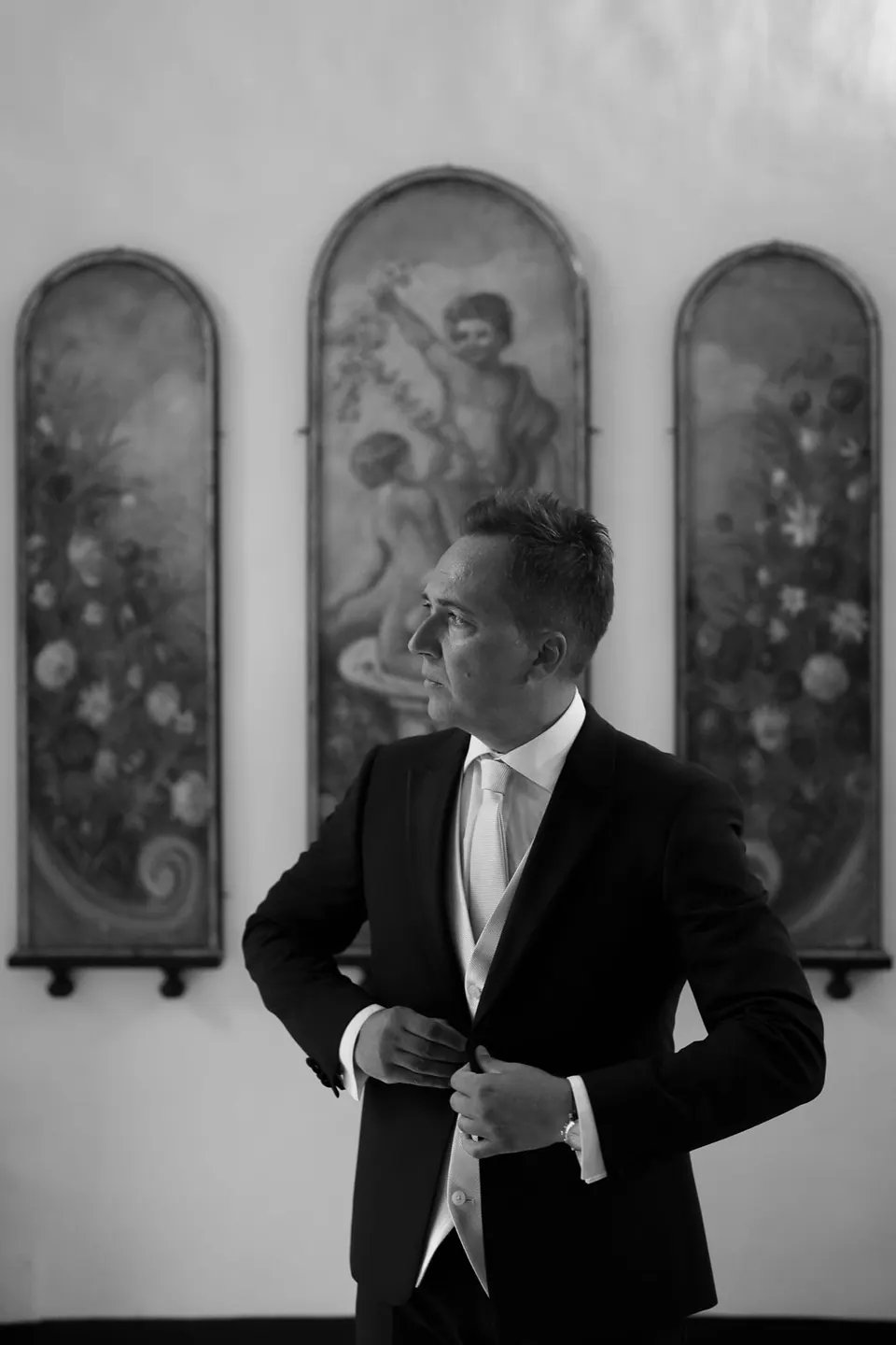 Black and white photo of the groom fastening his cuff in front of painted panels