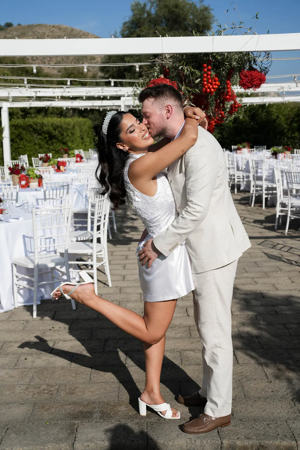 Le marié embrasse la mariée qui lève un pied, entre les tables et chaises blanches de la terrasse.
