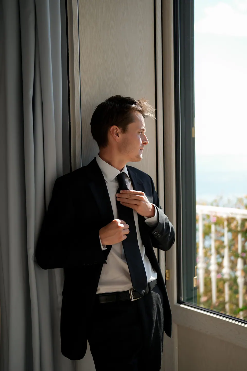Groom adjusting his tie beside a window.