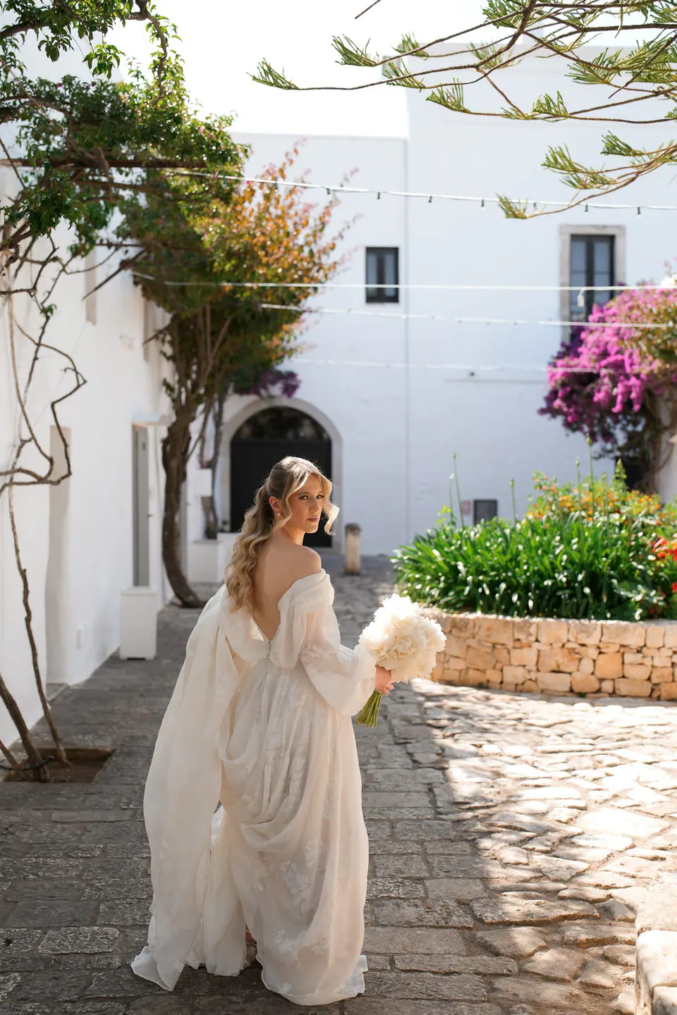 La novia junto a los muros blancos de la masseria, con flores al lado.