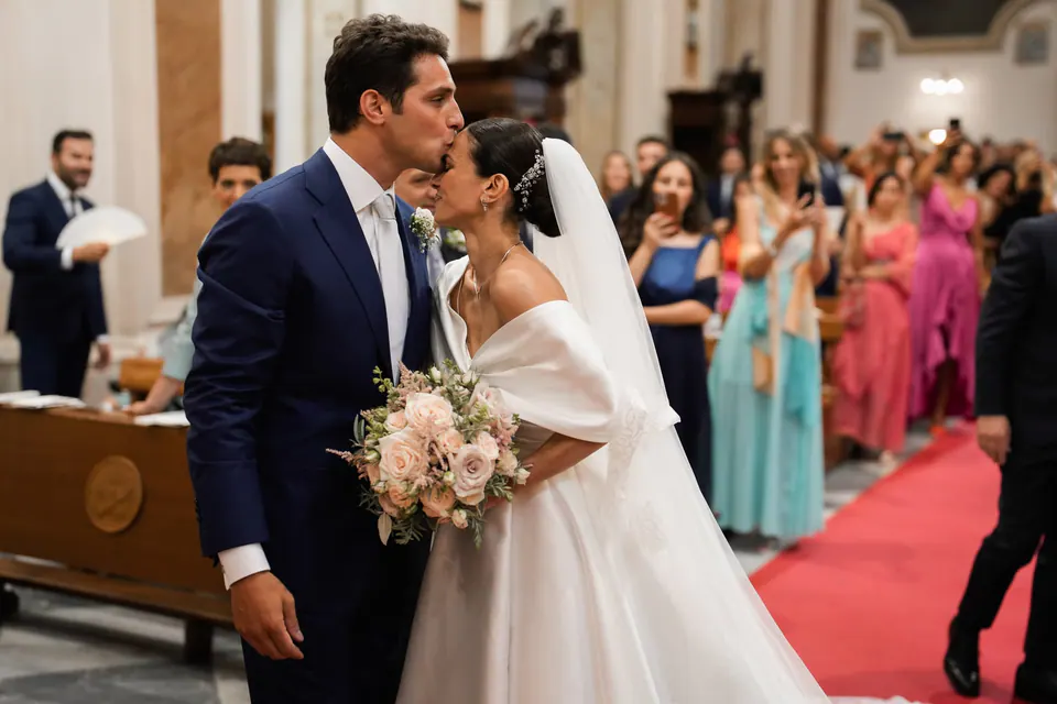 Lo sposo bacia la sposa all'altare, damigelle in abiti colorati alle spalle.