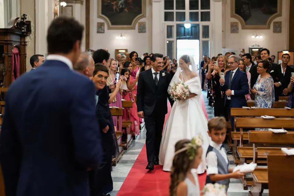 Sposa lungo il tappeto rosso della navata, lo sposo che attende in primo piano.