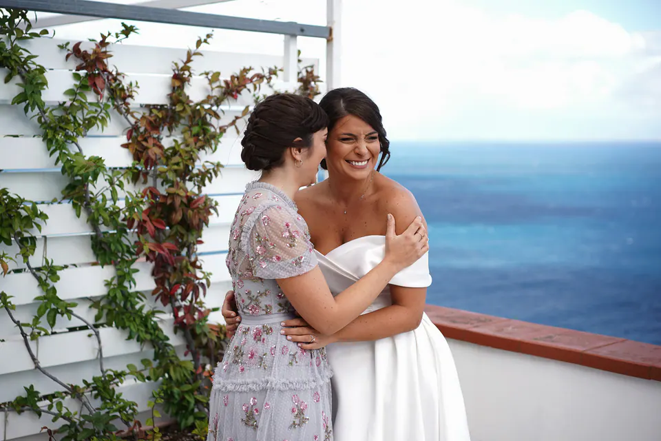 Bride embracing a guest in lilac lace on the terrace above the sea.