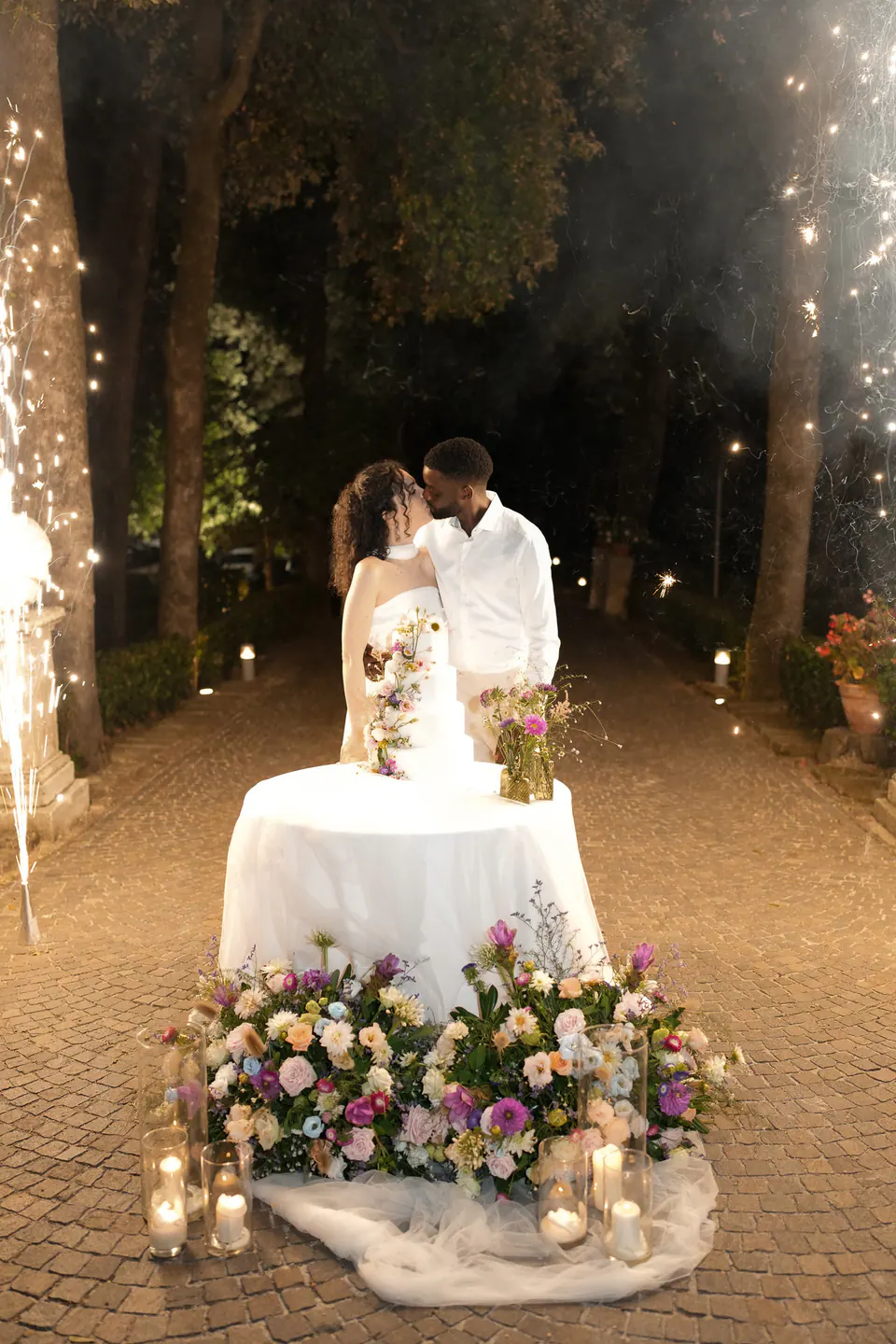 Mesa de la tarta bajo las guirnaldas en el jardín de noche, los novios cortando la tarta tras un lecho de flores.