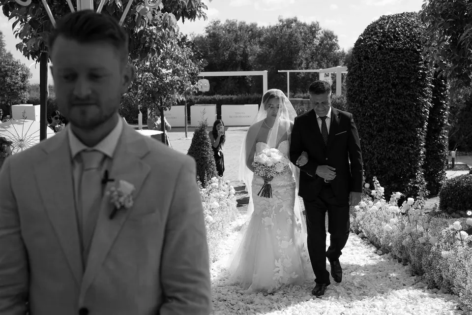 Le marié attend au premier plan tandis que la mariée remonte l’allée bordée de fleurs.