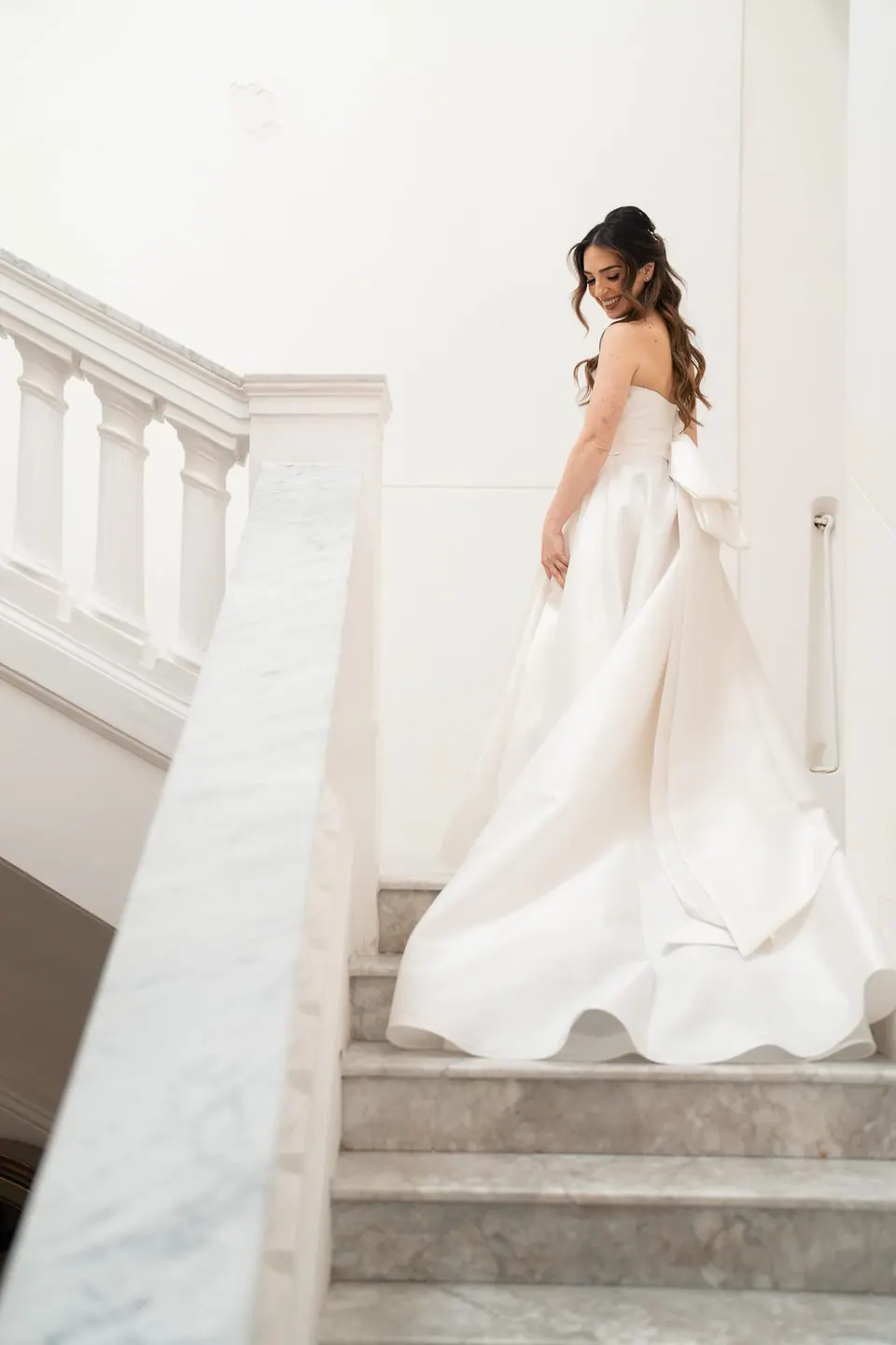 La novia baja una escalera de mármol blanco sujetando el vestido.