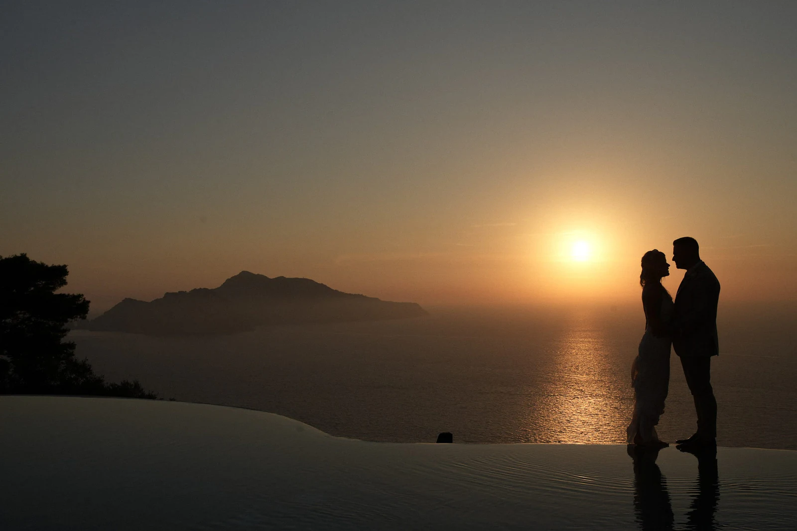 Sposi in controluce al tramonto accanto alla piscina a sfioro, Capri all'orizzonte.