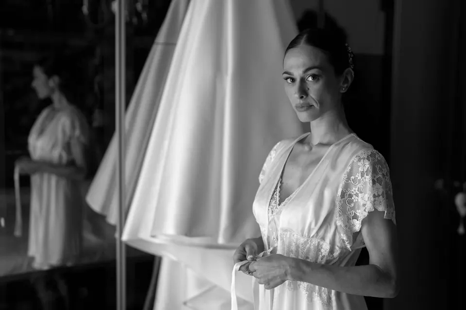 Foto in bianco e nero della sposa in vestaglia accanto all'abito appeso.