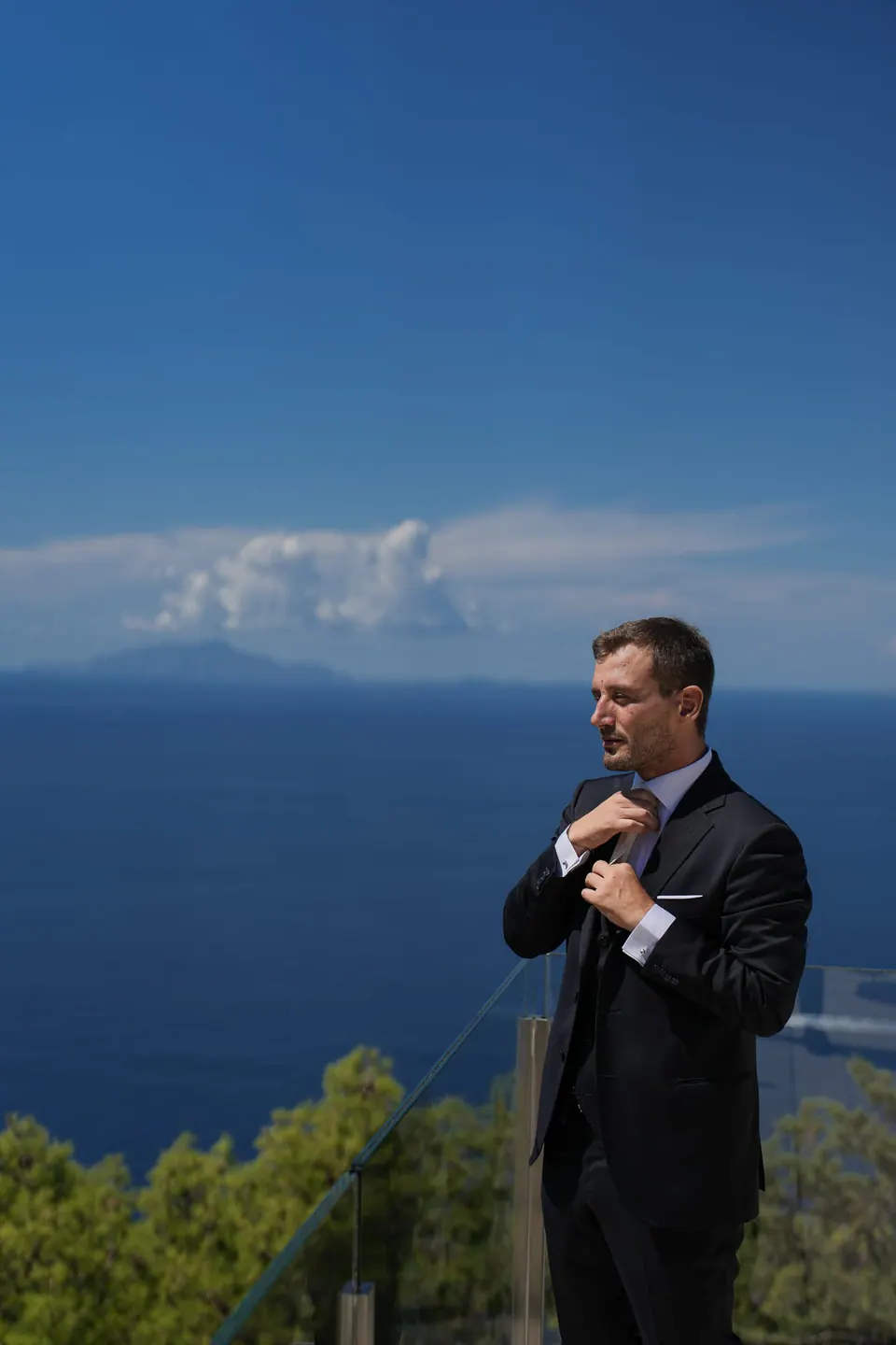 Lo sposo si sistema la giacca in terrazza, il mare blu intenso alle spalle.