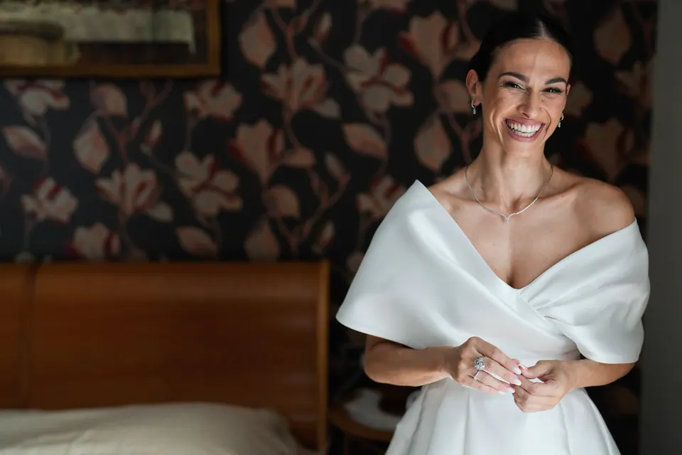 Sposa che ride con l'abito a spalle scoperte contro una carta da parati scura a fiori.