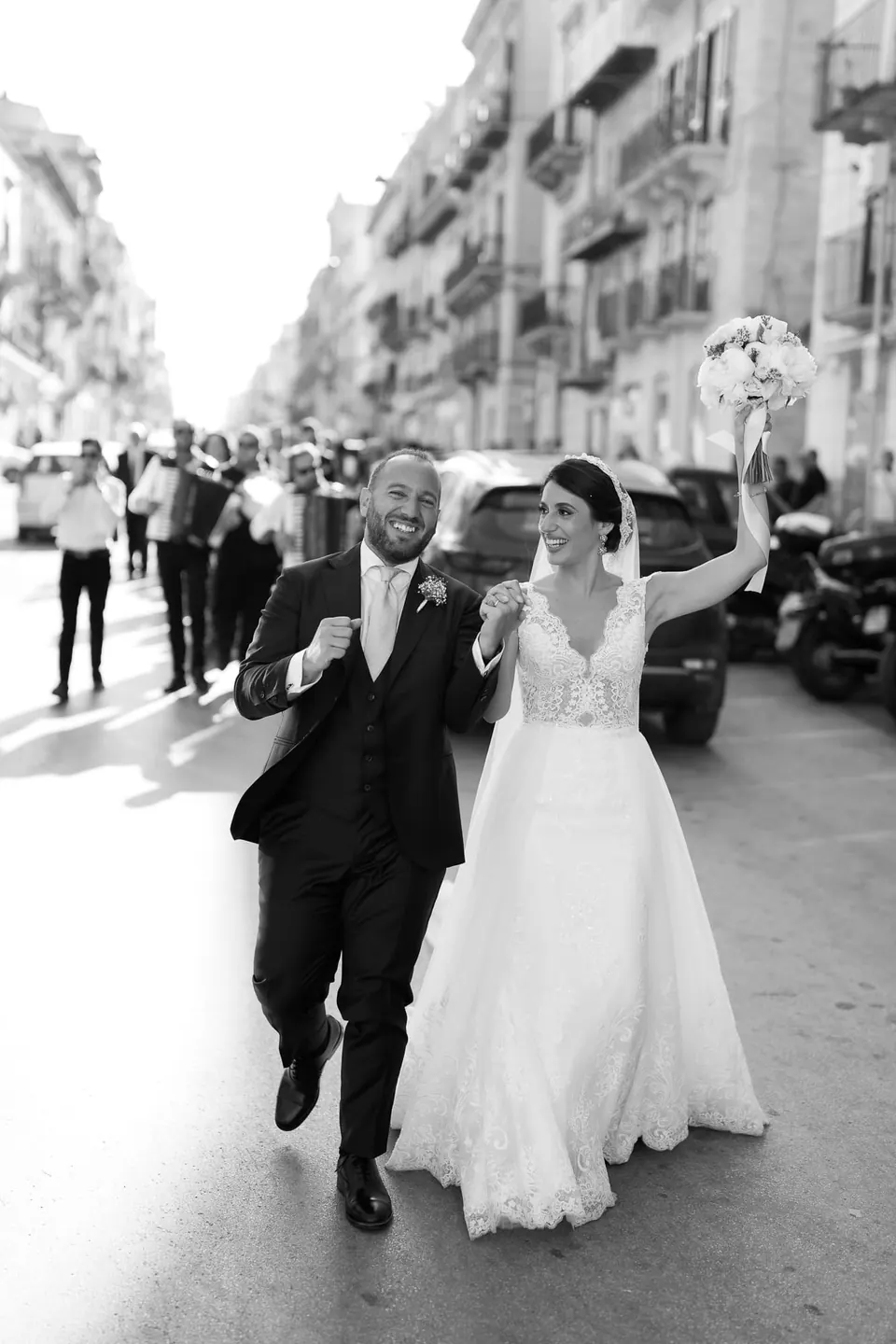 Black and white photo of bride and groom walking through a town street, the bride waving