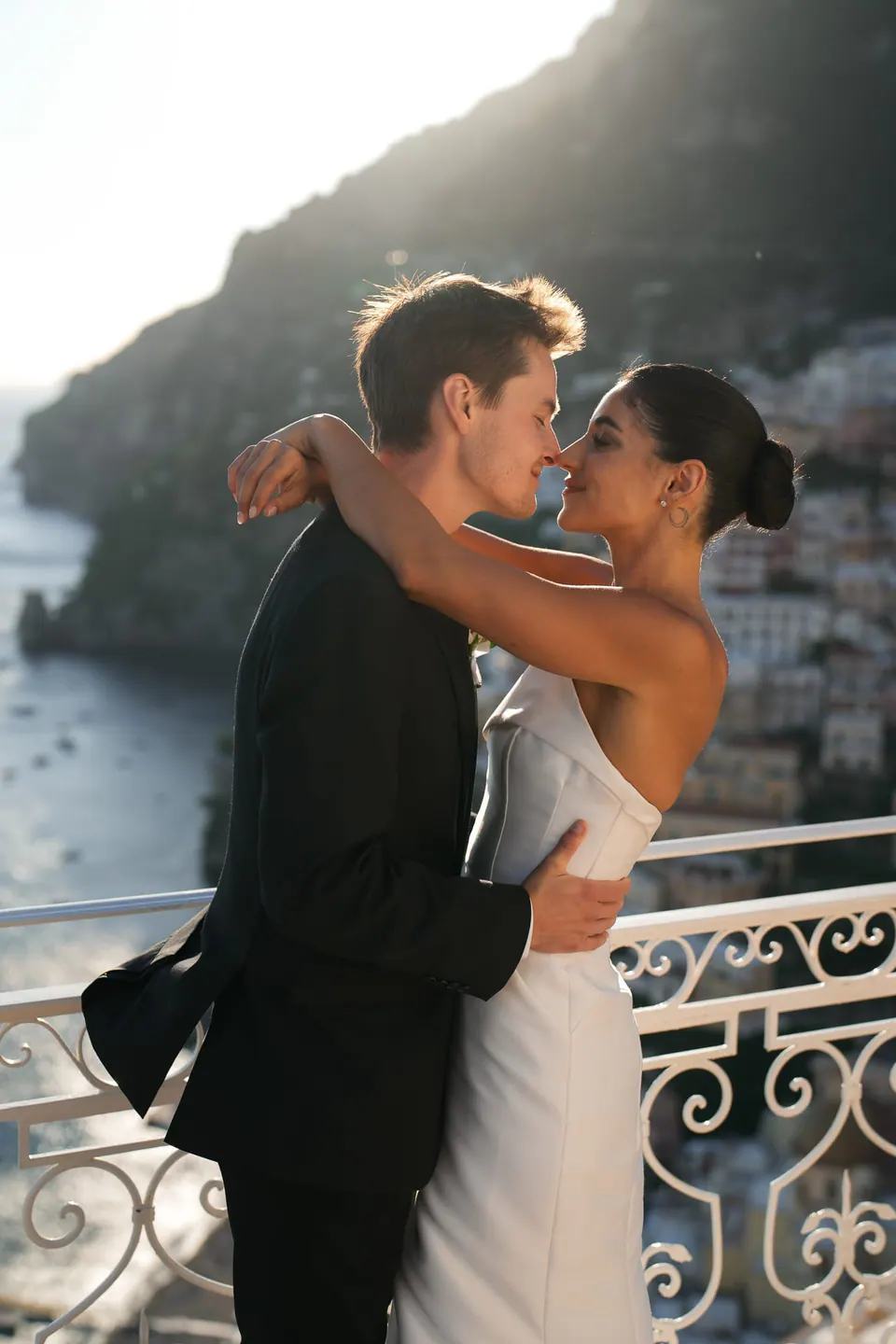 Bride and groom embracing at the railing, the coast of Positano behind.
