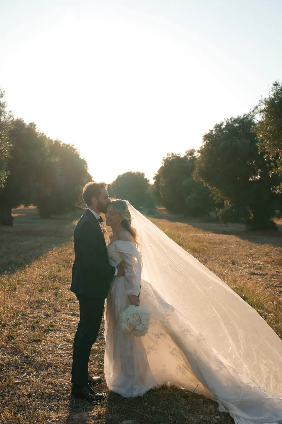 Los novios se besan en un olivar al atardecer, con el velo al viento.