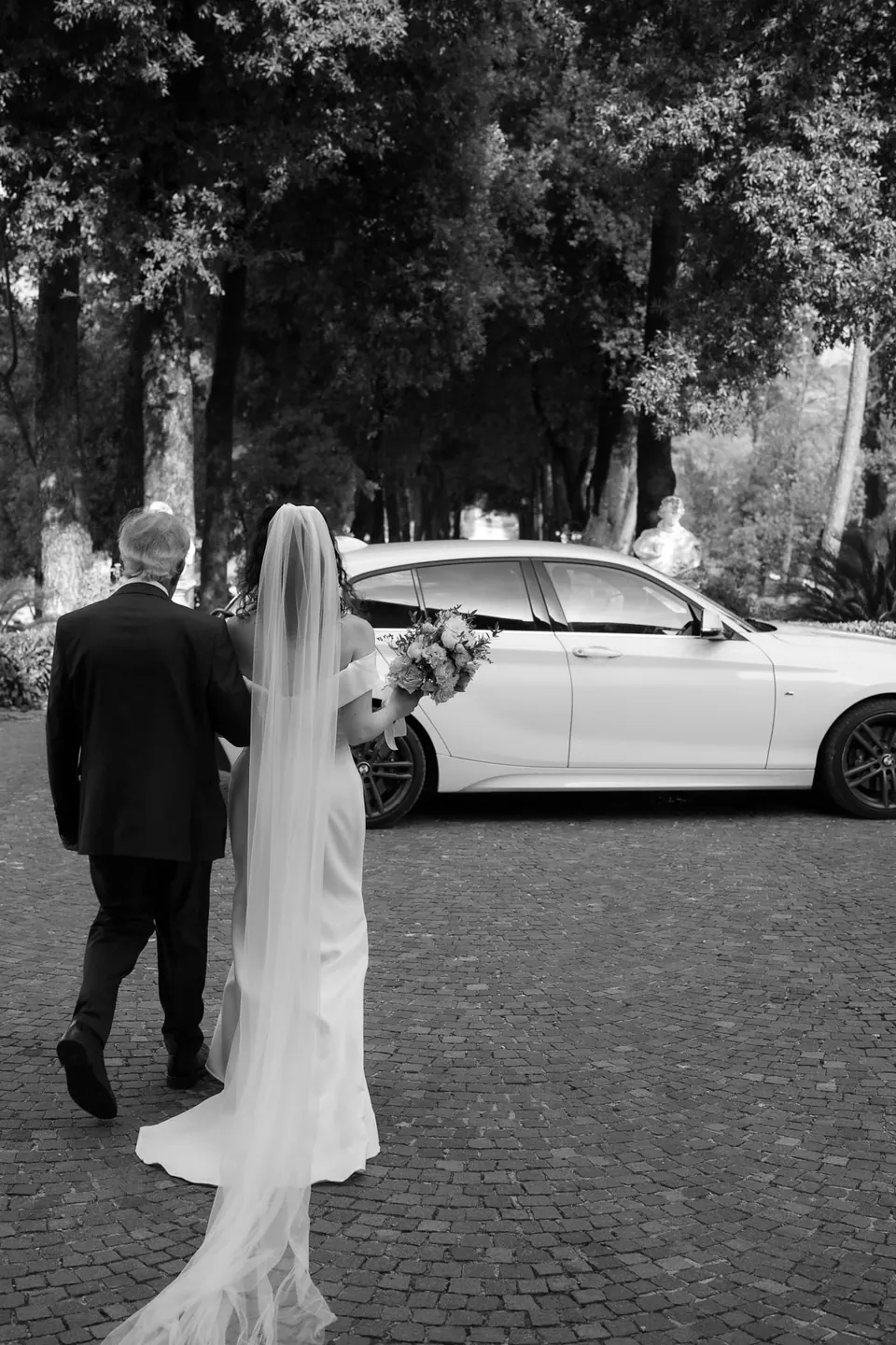 Foto en blanco y negro de la novia caminando hacia el coche blanco, con el velo arrastrando por el empedrado.