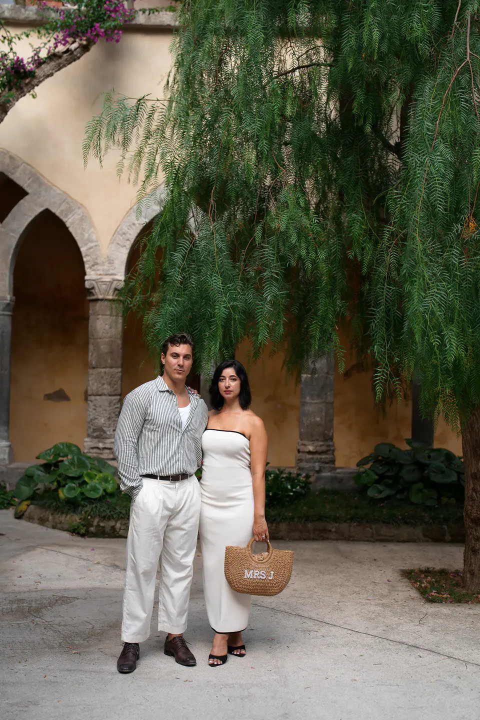 Couple standing in a cloister garden, a willow tree behind them.