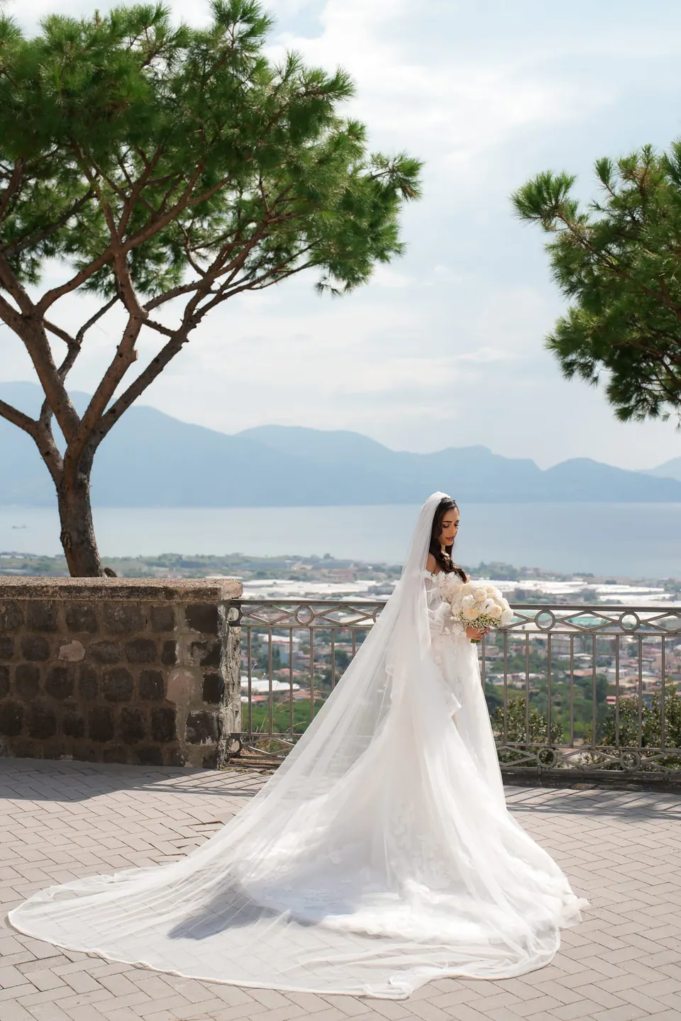 Bride on the terrace with her long veil spread on the stone, umbrella pines and the gulf behind.