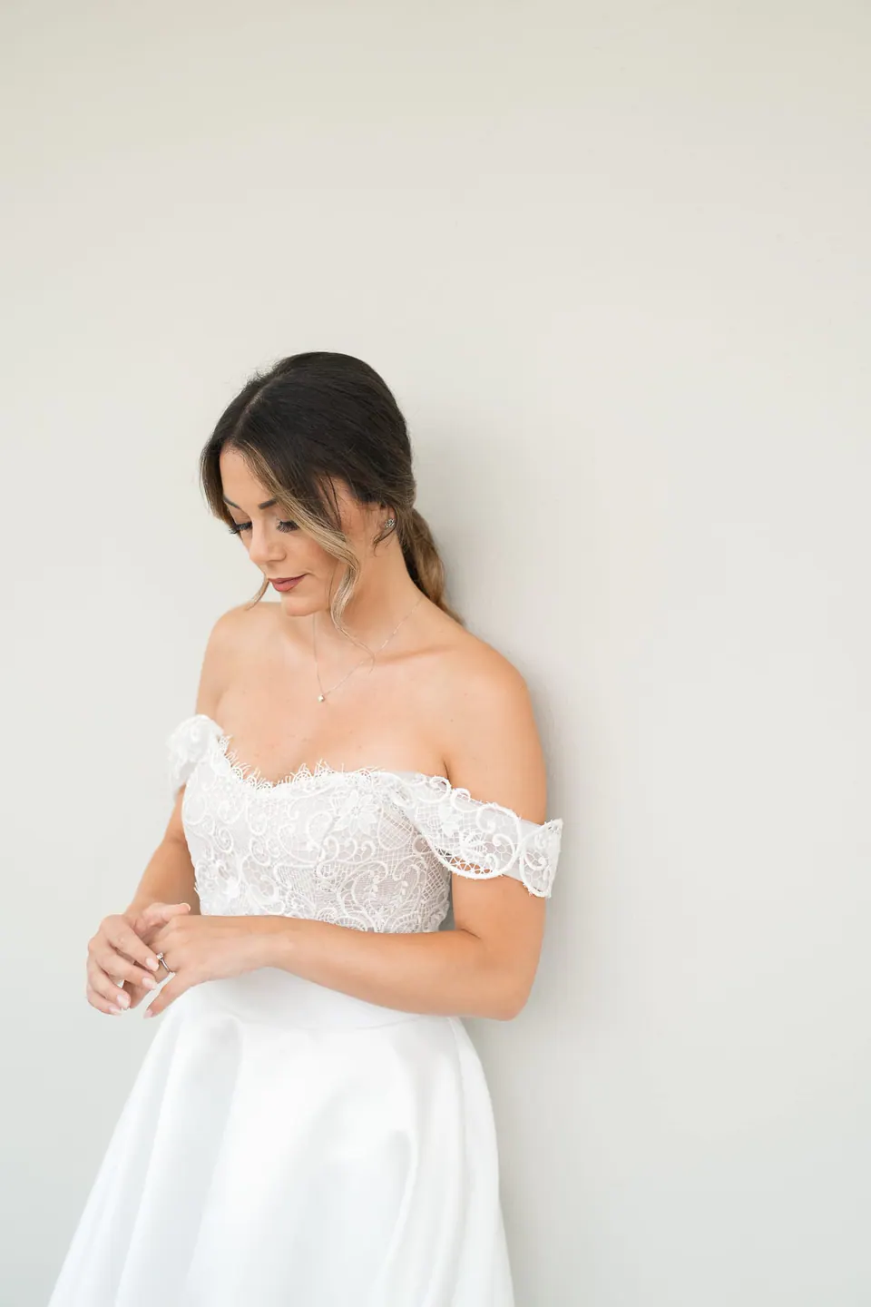 Bride in an off-the-shoulder lace dress against a white wall