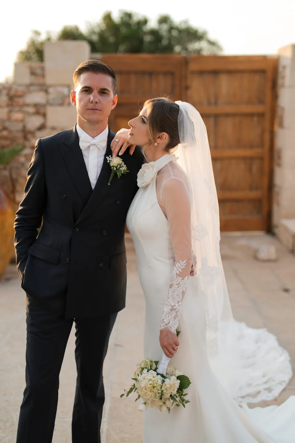 La sposa appoggiata alla spalla dello sposo, con muretti a secco e portone in legno alle spalle.