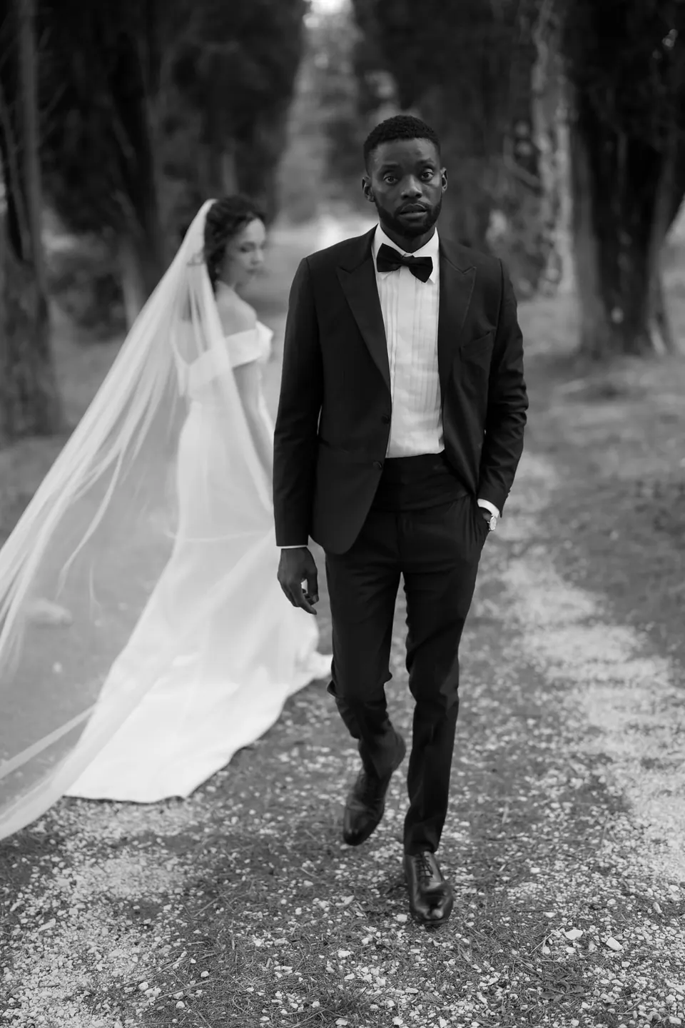 Foto en blanco y negro de los novios bajando por la avenida de cipreses, con el velo largo arrastrando.