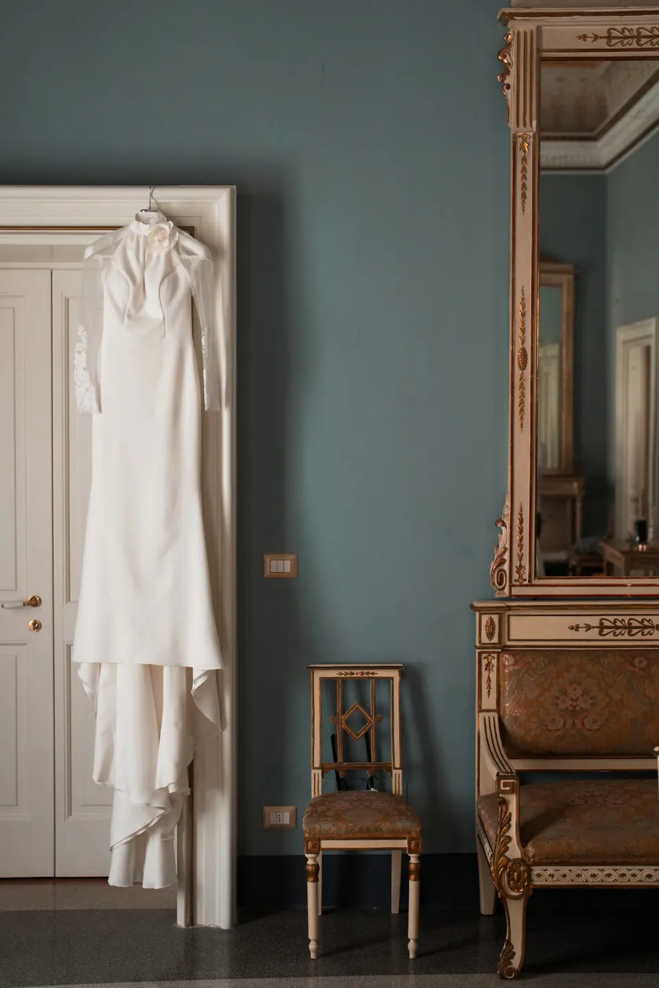 L'abito da sposa appeso a una porta bianca in una stanza con pareti celesti e mobili antichi dorati.