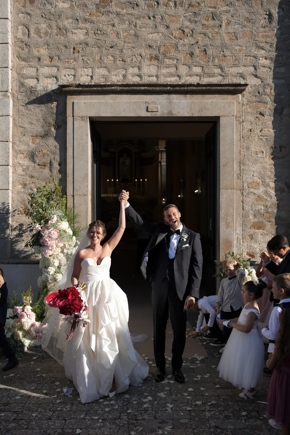 La sposa alza il bouquet all'uscita dalla chiesa, invitati e bambini intorno.