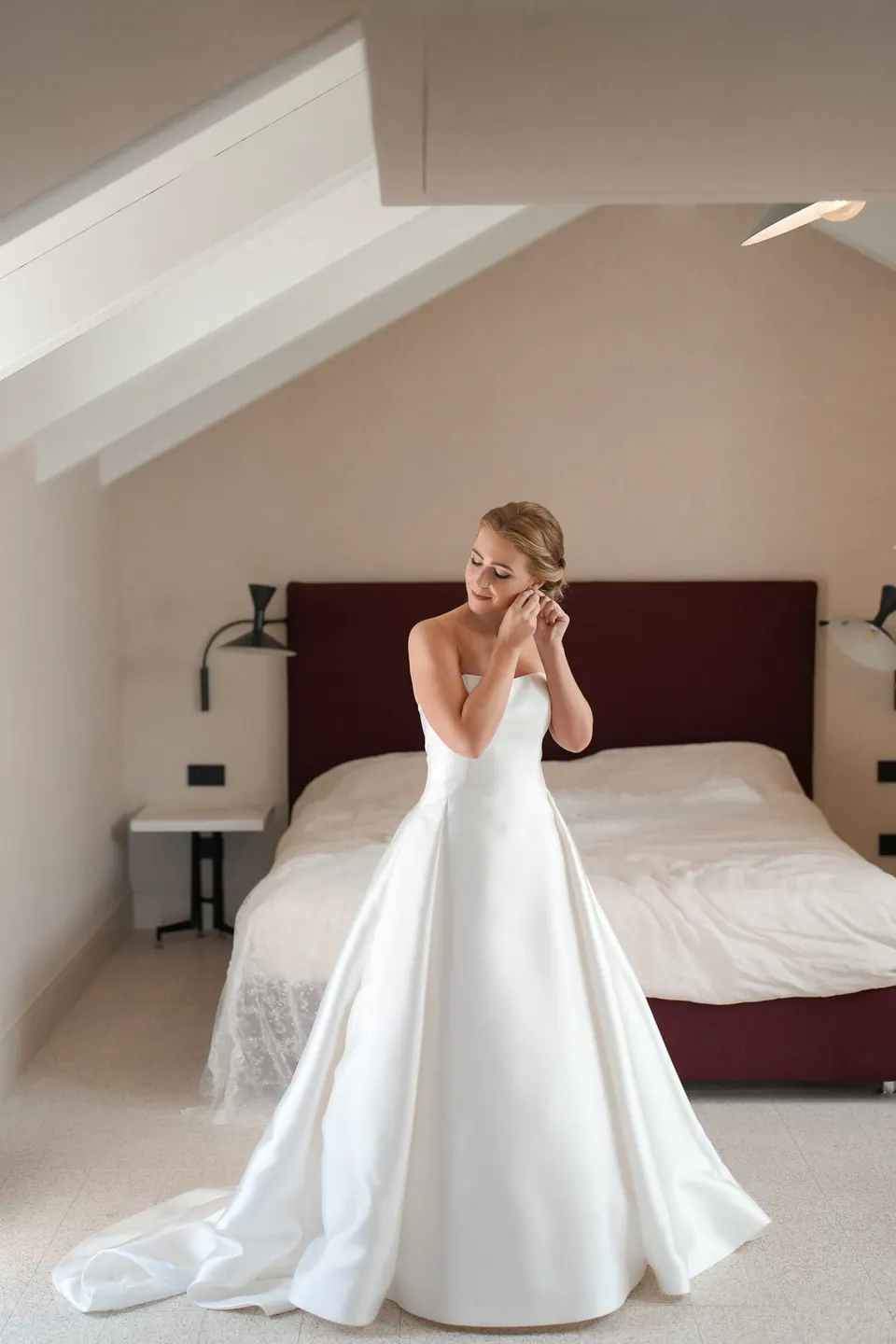 Die Braut legt im Dachzimmer die Ohrringe an, ihr Kleid liegt auf dem Boden ausgebreitet.