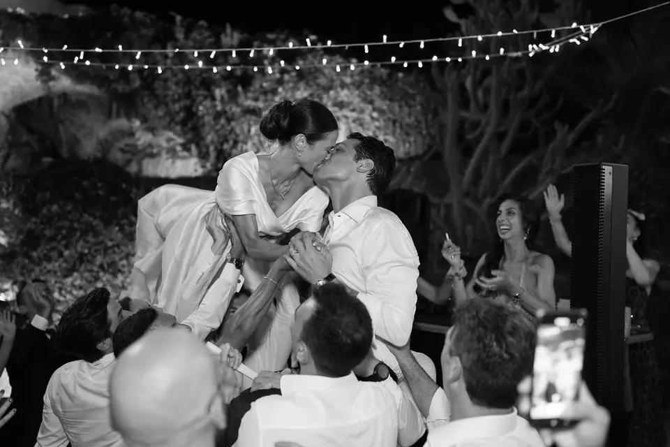Foto in bianco e nero del bacio degli sposi mentre gli invitati li sollevano alla festa.