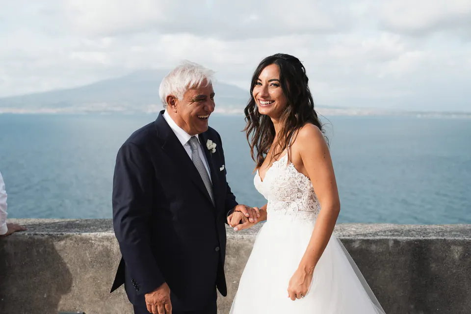Il papà e la sposa ridono in terrazza, il Vesuvio e il Golfo di Napoli alle spalle.