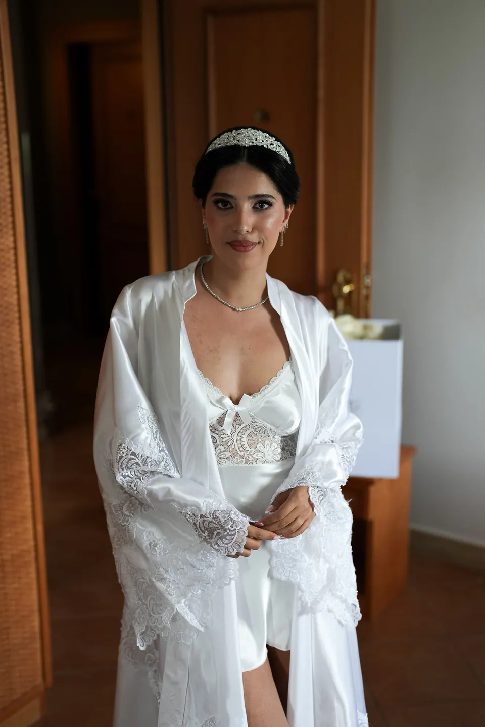 La mariée en peignoir bordé de dentelle et diadème sur le seuil de sa chambre.