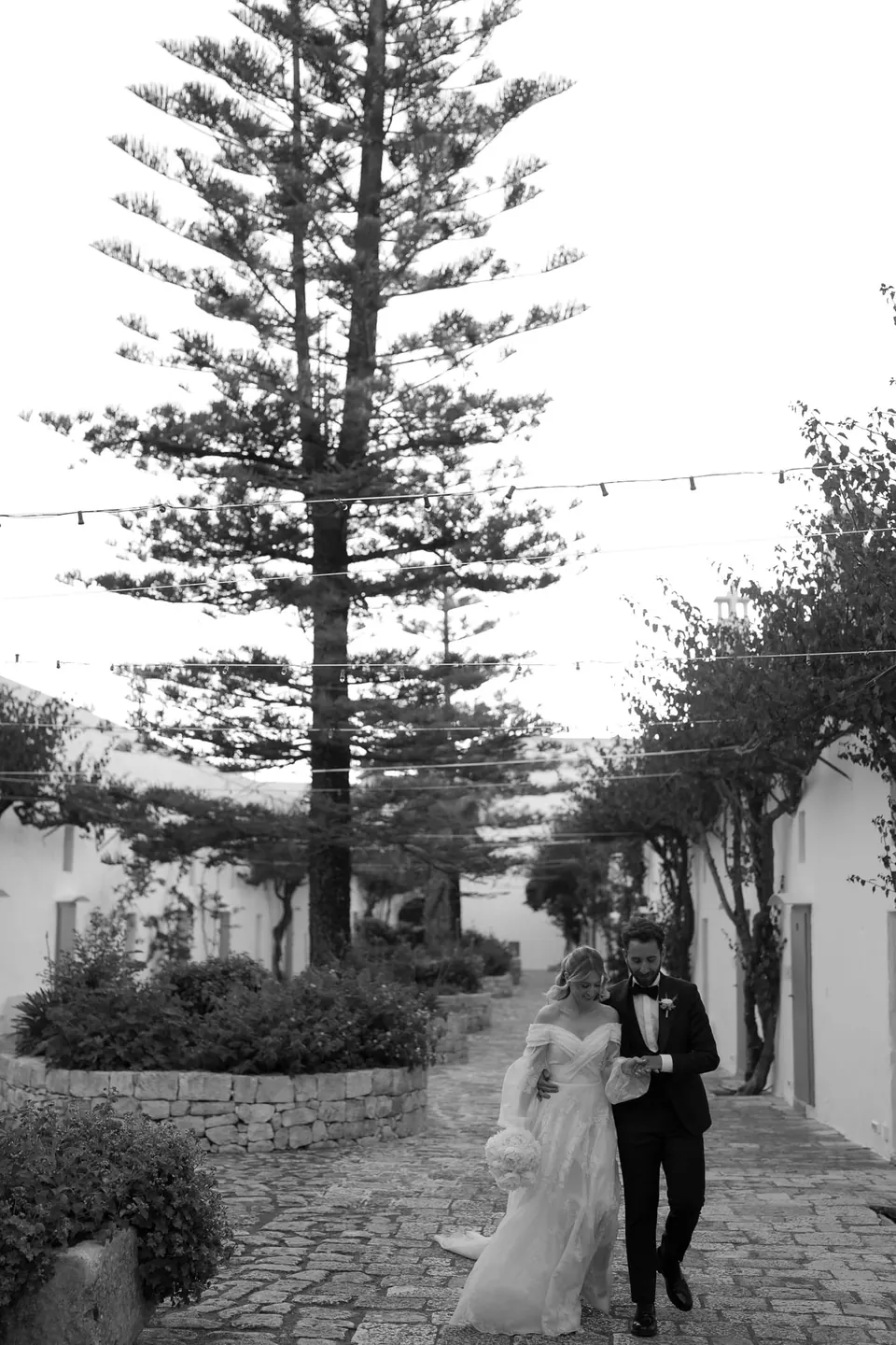 Los novios pasan junto a un pino alto en un pueblo blanco.