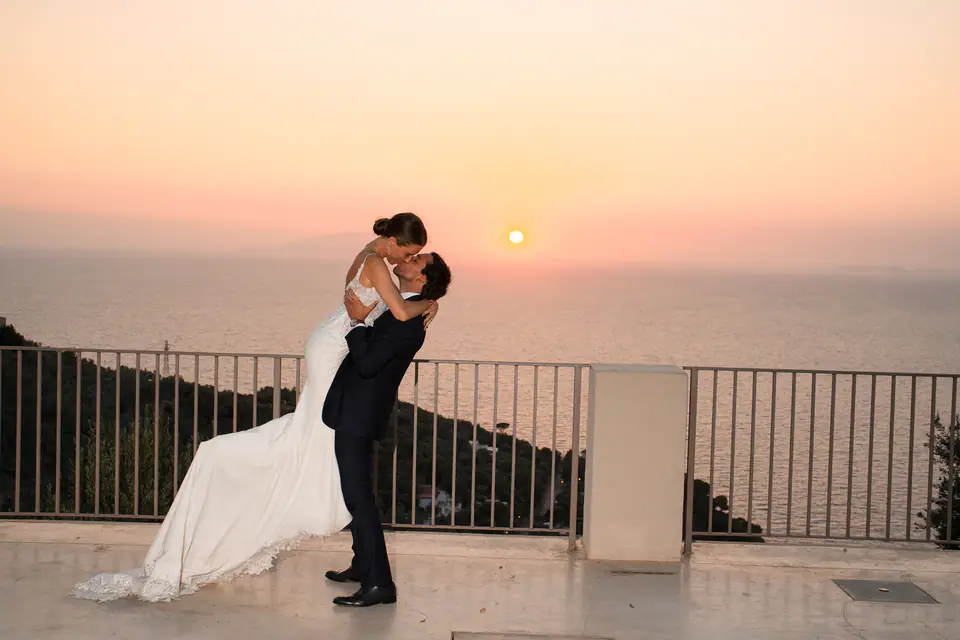 Groom lifting the bride at a terrace railing, the sun setting over the sea.